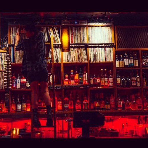 A bartender organizes vinyl records on a shelf above a well-stocked bar, illuminated by warm red lighting.