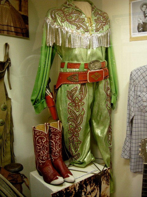A green and silver fringed cowboy outfit displayed on a mannequin, with red cowboy boots beside it.