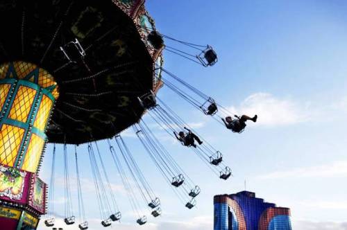 A colorful swing ride at an amusement park, with riders soaring against a blue sky and modern buildings in the background.
