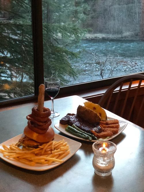 A hearty meal featuring a towering burger with onion rings, fries, and a side of steak, with a glass of wine by a river view.
