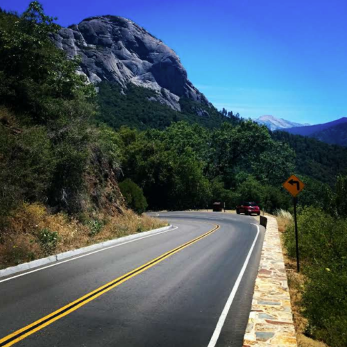 A winding road curves through a mountainous landscape under a clear blue sky, with greenery on either side.