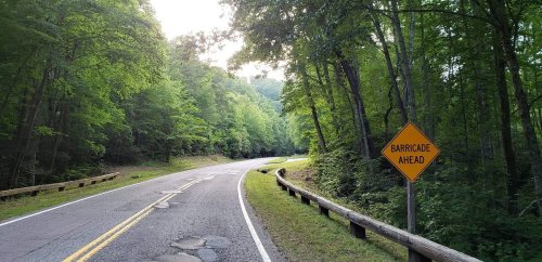 A winding road through lush green trees, with a warning sign that reads "BARRICADE AHEAD."