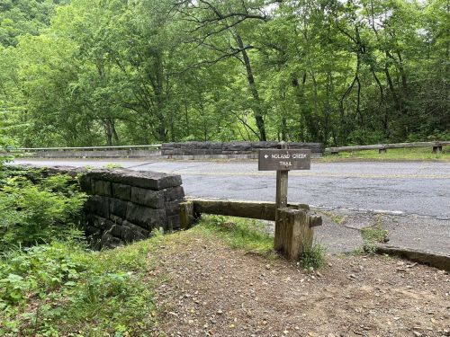 A wooden sign for the Moland Creek Trail near a stone bridge, surrounded by lush green trees and a paved road.