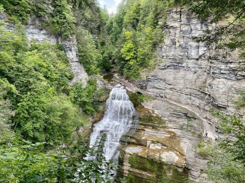 A scenic waterfall cascading down rocky cliffs, surrounded by lush green trees and a winding path along the water.