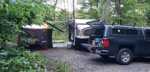 A camping setup with a pop-up trailer, a screened tent, and a truck parked in a wooded area.