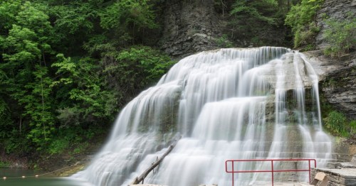A serene waterfall cascades over rocky cliffs, surrounded by lush green trees and a peaceful natural setting.