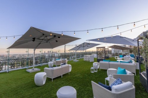 Rooftop lounge with white furniture, umbrellas, and string lights, overlooking a city skyline at sunset.