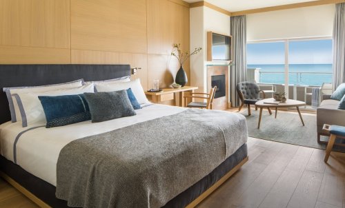 A modern hotel room with a king bed, ocean view, wooden accents, and cozy seating area.