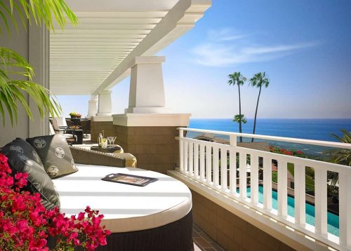 A sunny balcony with a lounge chair, flowers, and a view of the ocean and palm trees in the background.