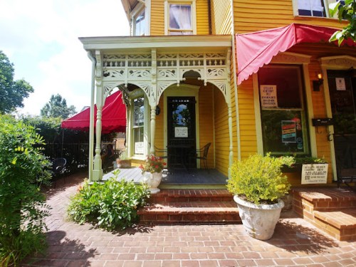 A charming yellow house with a decorative porch, red awnings, and potted plants on a brick pathway.