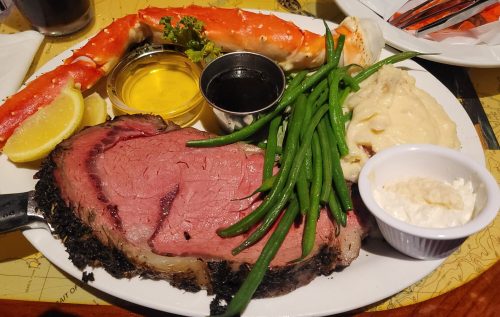 A plate featuring prime rib, crab legs, green beans, mashed potatoes, and sauces, garnished with lemon and parsley.