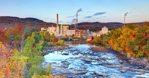 A scenic view of a factory by a river, surrounded by colorful autumn foliage and mountains in the background.
