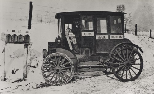 A vintage mail truck parked by a snow-covered roadside mailbox, with a driver visible inside.