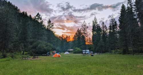 A serene campsite at sunset, featuring tents, picnic tables, and surrounded by tall trees and a grassy area.