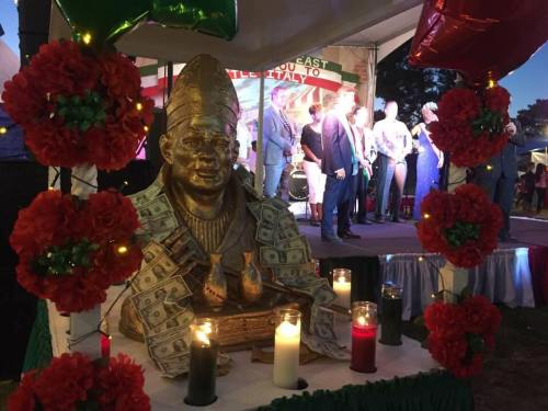 A statue draped in money sits in front of a stage decorated with flowers and lights during a festive event.