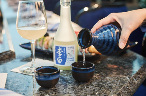 A hand pours sake from a blue ceramic bottle into two small cups, with a glass of drink and sushi in the background.