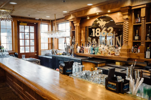 A wooden bar with a variety of bottles and glasses, featuring a sign that reads "1862 Saloon." Warm, inviting atmosphere.