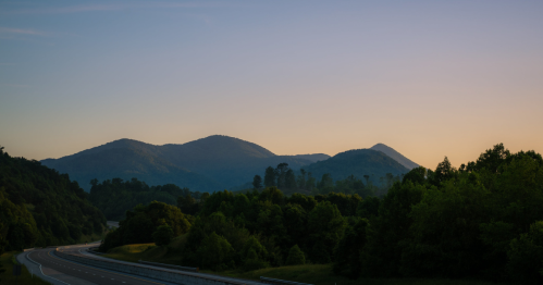 A serene landscape featuring rolling mountains at dusk, with a highway winding through lush greenery in the foreground.