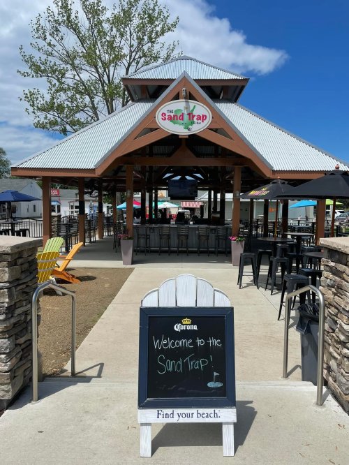 A welcoming sign at the entrance of a beach-themed venue called "The Sand Trap," with outdoor seating and umbrellas.