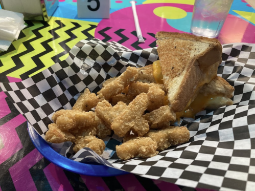 A plate with a grilled cheese sandwich and crispy, golden-brown fried snacks on a colorful, zigzag-patterned table.