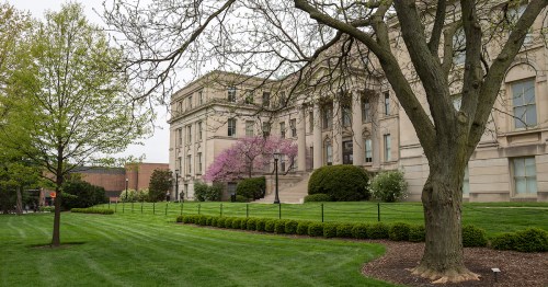 A landscaped area featuring green grass, trees, and a historic building with blooming pink flowers in the background.