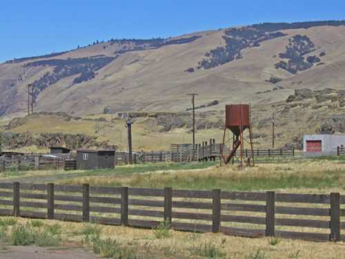 A rural landscape featuring rolling hills, a water tower, and wooden fencing under a clear blue sky.