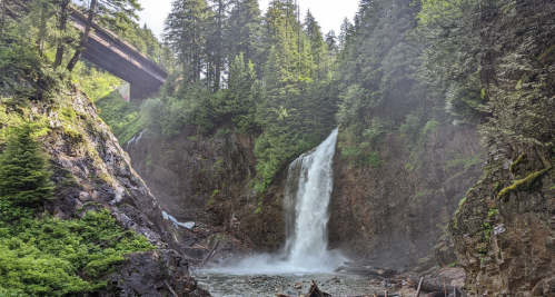 A waterfall cascades down rocky cliffs, surrounded by lush green trees, with a bridge visible above.