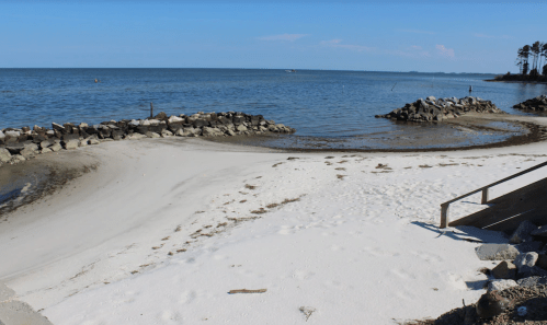 A serene beach scene with white sand, calm water, and rocky formations under a clear blue sky.
