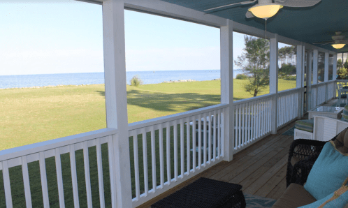 A spacious porch with white railings overlooking a green lawn and calm water in the distance under a clear sky.