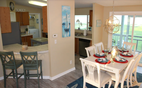 Cozy dining area with a table set for four, open kitchen, and a view of greenery through large windows.