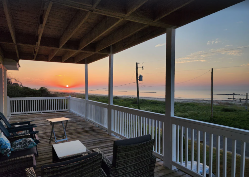 A serene sunset view from a porch overlooking the beach, with chairs and a table on a wooden deck.