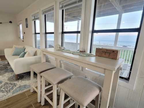 Bright coastal interior with a white sofa, bar stools, and large windows showcasing a scenic ocean view.