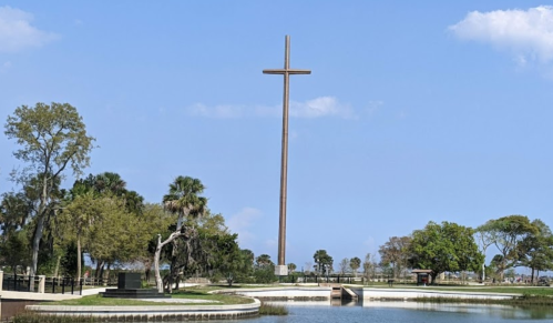 A tall wooden cross stands near a calm waterway, surrounded by trees and under a blue sky with scattered clouds.