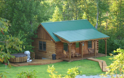 A cozy wooden cabin with a green metal roof, surrounded by trees, featuring a porch and a hot tub on the deck.