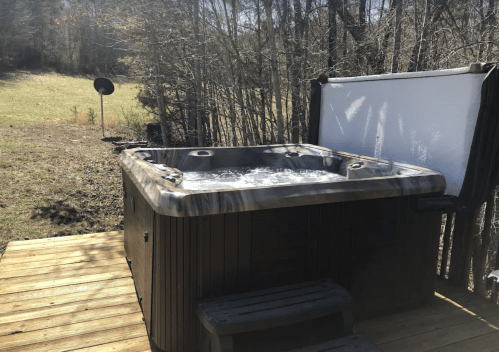 A hot tub on a wooden deck surrounded by trees, with a grassy area and a white screen in the background.