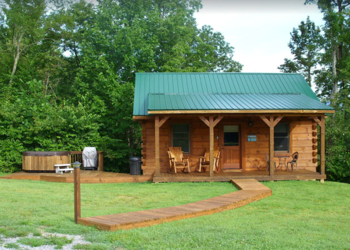 A cozy wooden cabin with a green metal roof, surrounded by trees, featuring a hot tub and a wooden deck.