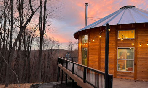 A round wooden cabin with a deck, surrounded by trees, under a colorful sunset sky. String lights illuminate the exterior.