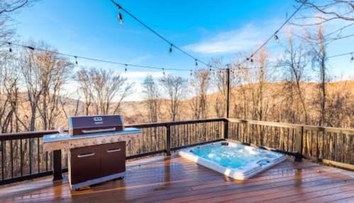 A wooden deck with a grill and hot tub, surrounded by trees and a clear blue sky.