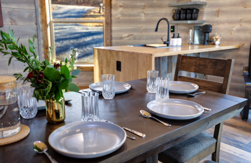 A rustic dining table set with plates and glasses, with a kitchen area visible in the background.