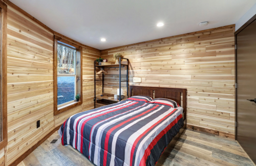 Cozy bedroom with a wooden accent wall, a striped bedspread, and a window overlooking a forested area.