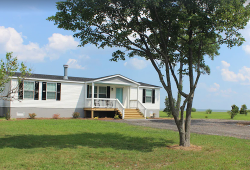 A white modular home with a front porch, surrounded by green grass and a tree, under a blue sky with clouds.