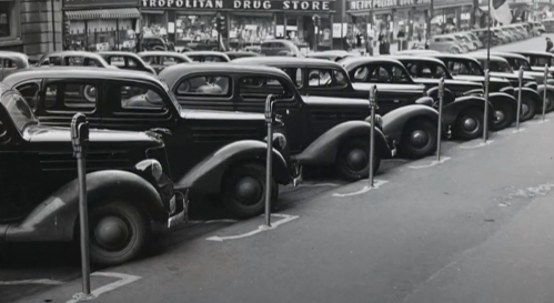 A row of vintage cars parked along a city street, with a drug store in the background. Black and white photo.