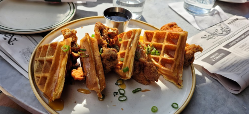 A plate of waffles and fried chicken, drizzled with syrup and garnished with green onions, served with a small cup of sauce.