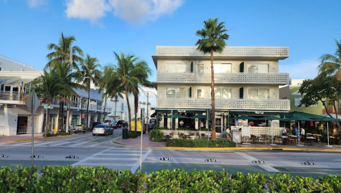 A sunny street scene featuring palm trees, a multi-story building, and outdoor dining areas in a vibrant urban setting.