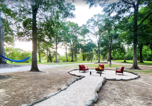 A cozy outdoor seating area with red chairs around a fire pit, surrounded by trees and a hammock in the background.