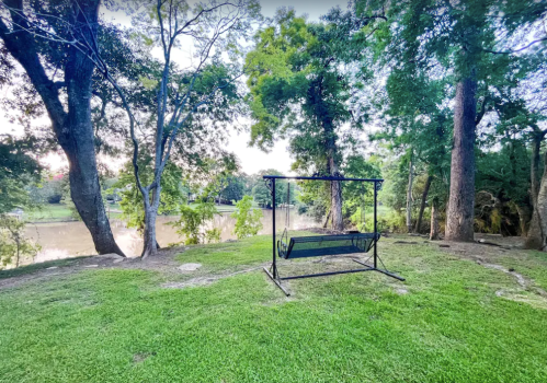 A swing set on a grassy area near a calm river, surrounded by lush green trees.