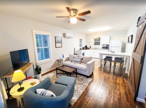 Cozy living room with a sofa, TV, and kitchen area featuring a dining table and modern appliances. Bright and inviting space.
