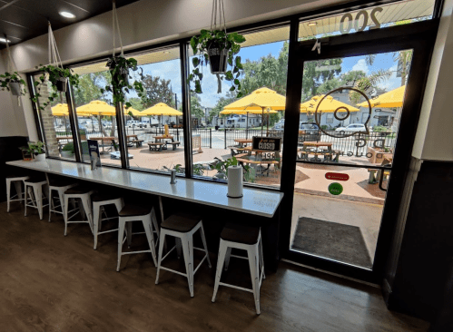 Bright café interior with a long counter, stools, and large windows overlooking an outdoor seating area with umbrellas.