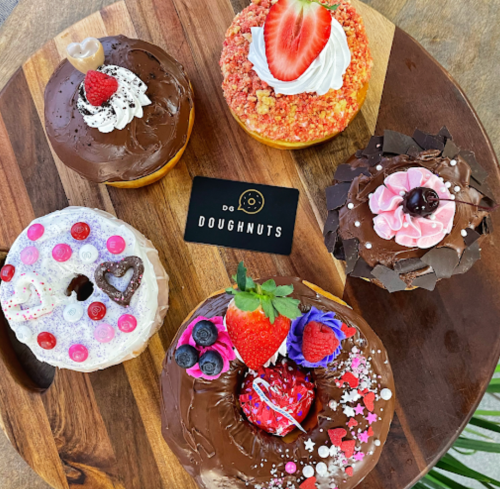 A wooden platter displays five colorful, decorated doughnuts alongside a "Do Doughnuts" business card.