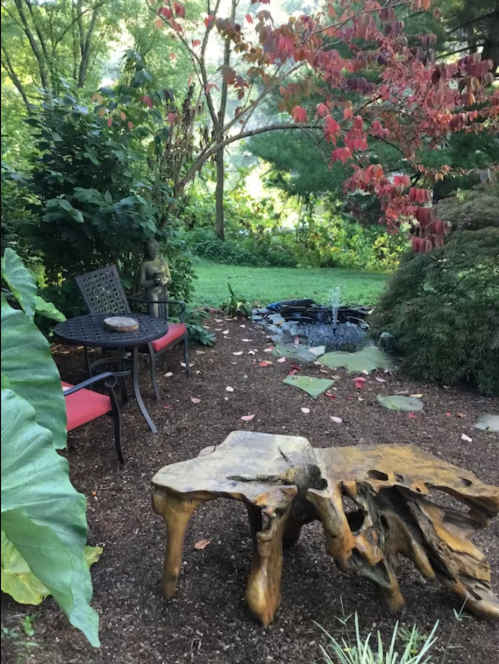 A serene garden scene featuring a wooden table, chairs, and a small pond surrounded by lush greenery and autumn leaves.
